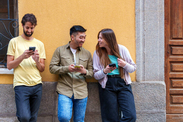 Захвачено яркое городское товарищество: трое разнообразных друзей, одетые в яркие городские наряды, погруженные в мобильный контент, прислонившись к городской стене.