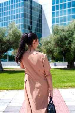 Beyaz bir yönetici model, canlı yeşilliklerle çevrili camdan gökdelenlerin finans bölgesi arkaplanına doğru ilerliyor..