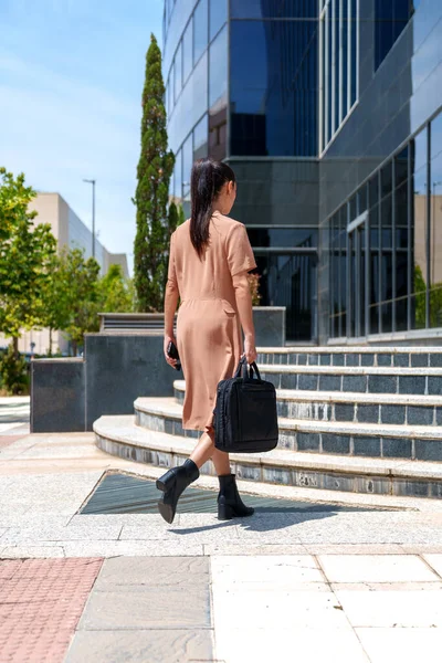 İş kıyafetleri içinde beyaz bir yönetici model, finans bölgesindeki ofisinin giriş merdivenlerine tırmanır, cam binalarla çevresini çevreleyen yeşil, ekolojik verimliliğe sahip.