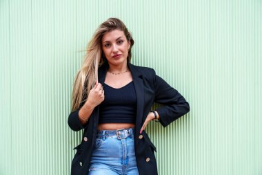 Çekici, sarışın, beyaz bir model, büyüleyici bir bakışla canlı yeşil bir duvara karşı duruyor. Zarafet ve kendine güven saçıyor..