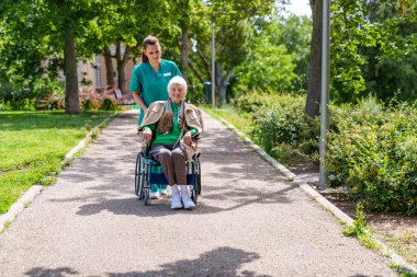 Neşeli bir şehir parkında güneşli bir gün, şefkatli bir hemşire yaşlı bir kadını yemyeşil bir yerde tekerlekli sandalyeye oturtuyor..