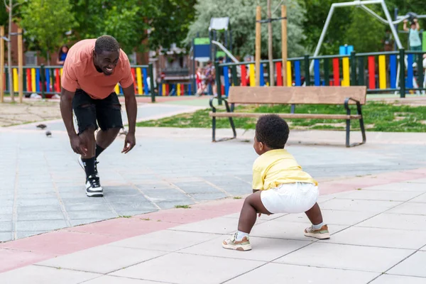 Happiness abounds as African dad and baby play in vibrant city park with lush green trees, beaming with joy.