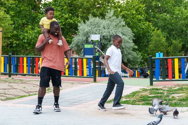 African dad with his two kids at a vibrant park. The elder playfully scares pigeons while the baby sits on his shoulders, pure happiness.