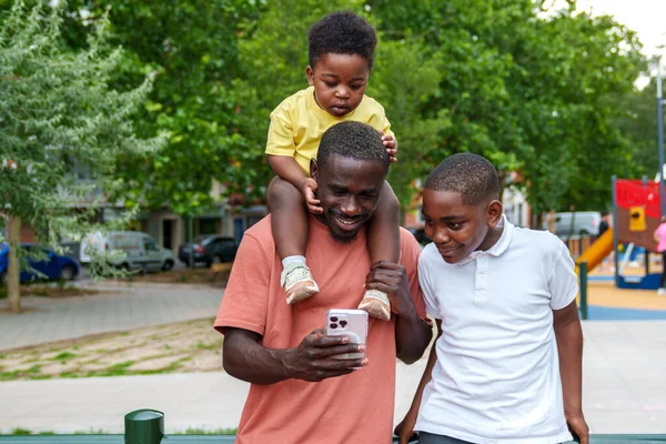 Happy African dad and kids in vibrant park; baby on shoulders, all smiles, captivated by mobile.