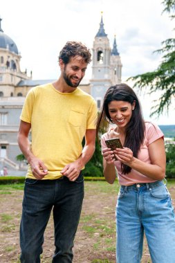 Parkta mutlu Kafkasyalı arkadaşlar, telefonlarını paylaşıyorlar, arka planda tarihi şehir anıtıyla..