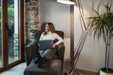 Evdeki kanepede bilgisayarı kullanan ve pencereden bakan genç bir kadın. Uzaktan evde çalışıyor. İnternet üzerinden kurslar alıp öğreniyorum..