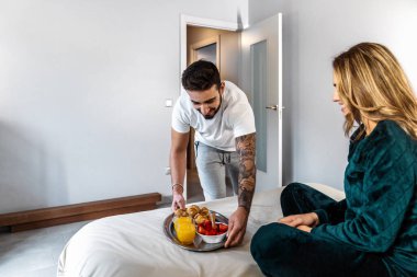 Pijamalı genç bir çiftin kocası onu yatakta bekleyen karısı için hazırladığı kahvaltıyı getiriyor. Koca, karısına sürpriz olsun diye kahvaltıyı yatakta bırakır. Romantik kahvaltı. Bir tepside kahvaltı. Yüksek kalite fotoğraf.