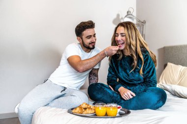 Pijamalı genç bir çift modern yatak odalarında yatakta sağlıklı kahvaltı yaparak harika vakit geçiriyorlar. Romantik kahvaltı. Yurt içi yaşam tarzı.