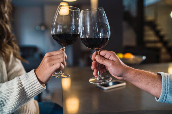 Close-up of the hands of an unrecognizable man and woman. Two young people with a glass of wine in a romantic and happy evening in the kitchen of their rural home. Couple toasting with a glass of red wine.