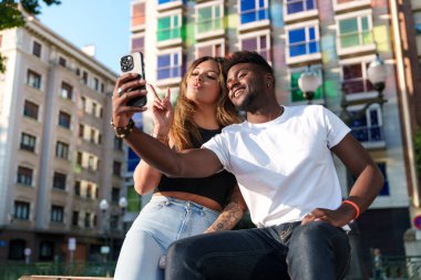 İki çekici, farklı şehir kıyafetleri giymiş model renkli şehir binalarına karşı neşeli bir selfie anı paylaşıyor..