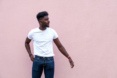 Beyaz tişörtlü ve kot pantolonlu genç Afrikalı erkek yumuşak pembe arka planda, düşünceli bir ifadeyle yana bakıyor..