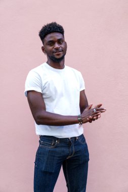 Beyaz gömlekli ve kotlu yakışıklı Afrikalı adam hoş bir gülümsemeyle poz veriyor, yumuşak pembe bir arka plana karşı sıradan bir güven yayıyor..