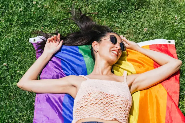 Joyful lady in sunglasses lies on LGBT pride flag on grass, enjoying summer.