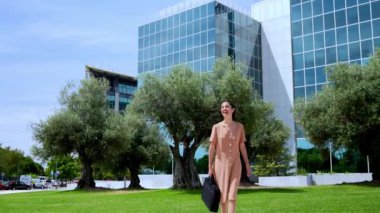 Şık giyinmiş şık bir iş kadını evrak çantasıyla yürüyor, 4K ağır çekimde modern cam bina parkında gülümsüyor..