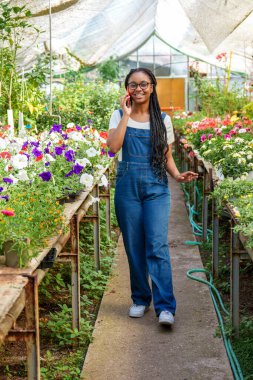 Neşeli Afrika kökenli kadın telefonda konuşuyor. Etrafı canlı sera bitkileriyle çevrili, günlük kot giyimli..