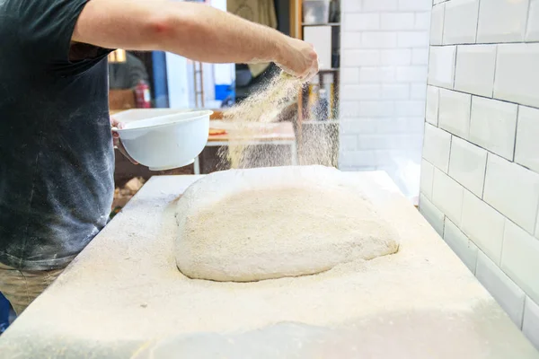 Baker dusting flour over bread dough on worktable in a bakery, with white tiles in the background.