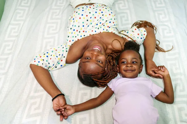 African mom with braids and polka dot top laughs with her child in a cozy bedroom setting.
