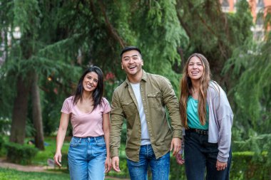 Neşeli üçlü yürüyüş yapmayı seviyor: İspanyol kadın, Asyalı-Çinli adam ve şehir parkındaki arkadaş.