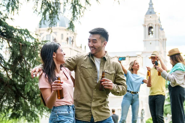 Cheerful mixed-ethnicity couple sharing laughs and beers in a vibrant city park, with historic building background.