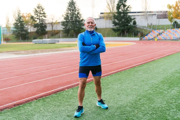 Smiling elderly athletic man in blue sportswear stands on a track, ready for his morning run