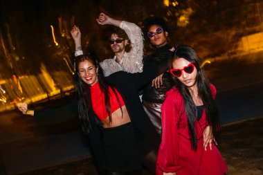 Dört neşeli çoklu etnik arkadaş gece ışıkları altında dans ediyor, mutluluk ve birliği ifade ediyorlar..