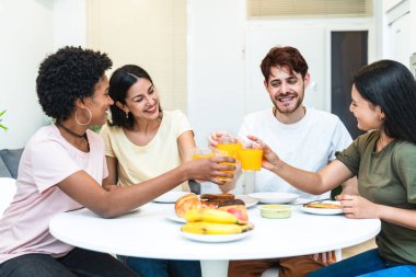 Dört çok ırklı arkadaş taze meyve suyu ve meyveli kahvaltıda gülüp kadeh kaldırıyor..