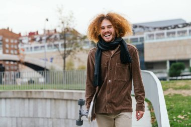 Dışarıda kaykaylı neşeli kızıl saçlı erkek. Kahverengi ceket giyiyor, şehir gününün tadını çıkarıyor..