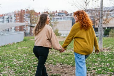 Neşeli kızıl saçlı ve esmer kadınlar, yaprakların arasında el ele, parkta rahatça yürüyorlar..