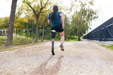 Protez bacaklı bir erkek koşucunun arka görüntüsü, spor kıyafetleriyle, yemyeşil bir parkta çakıl yolunda koşuyor..