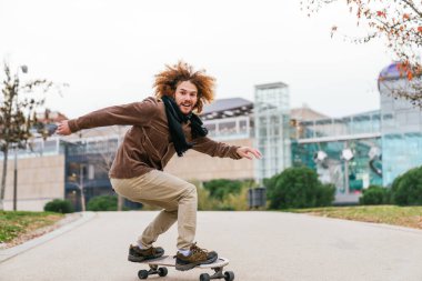 Şehir parkında afro saçlı kaykaylı genç, beyaz bir adam. Kahverengi ceket, atkı, sonbaharın tadını çıkarıyor..