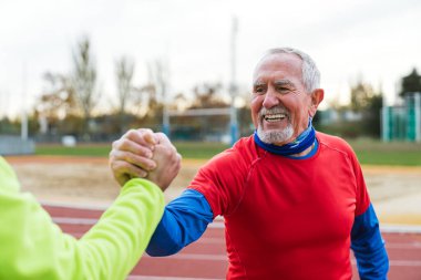 Kırmızı tişörtlü, beyaz sakallı neşeli yaşlı bir adam koşu yolunda yeşil gömlekli bir akranla el sıkışıyor..