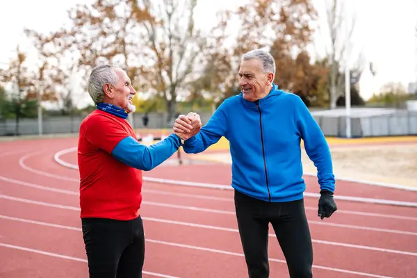 Two joyful senior male runners share a handshake on a sports track, exhibiting companionship and healthy lifestyle.
