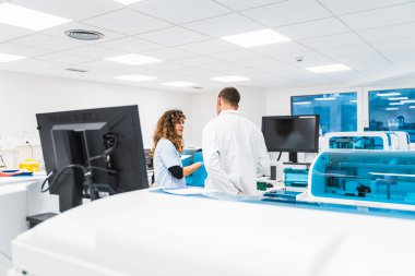 İki laboratuvar teknisyeni, temiz ve parlak bir hastane laboratuvarında kan analiz makinelerinin arasında sohbet ediyorlar..