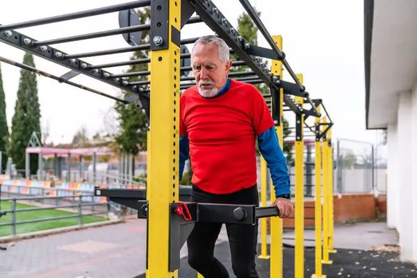 Focused elderly man in red exercising with determination on monkey bars at a public park, showcasing strength and agility