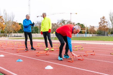 Takım arkadaşlarıyla spor kıyafetlerini tartışırken kırmızı kıyafetli yaşlı sporcu çeviklik ekipmanlarını düzenliyor.