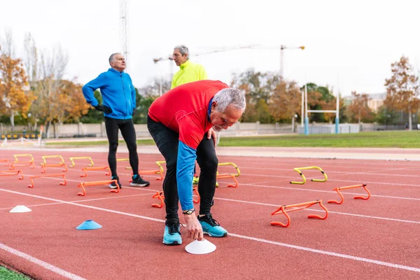 Mature runners on a track field, one stretching, others setting training hurdles for a workout.