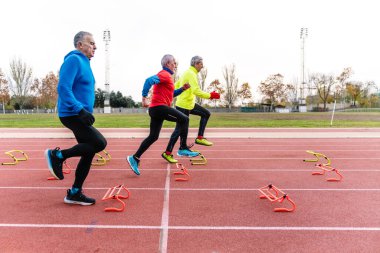 Bir grup yaşlı adam atletizm pistinde renkli engellerle çeviklik eğitimi alıyor.