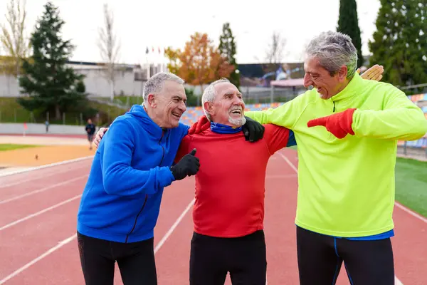 Group of joyful elderly friends in sportswear share a laugh and friendly gestures post-run on an outdoor athletic track.