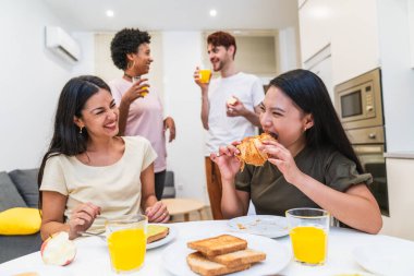 İki kadın, arkadaşlarıyla güneşli bir kahvaltı masasında kruvasan ve tost yerken içten bir kahkahadan zevk alır..