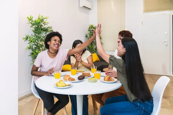 Friends enjoy a lively breakfast moment, with two women high-fiving over a table laden with fresh juice and baked goods.