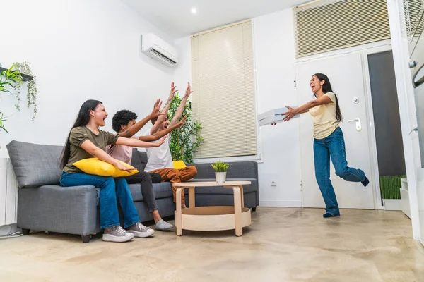 Woman joyfully entering a living room with pizza as friends cheer from the couch. A happy scene of friendship and food sharing.