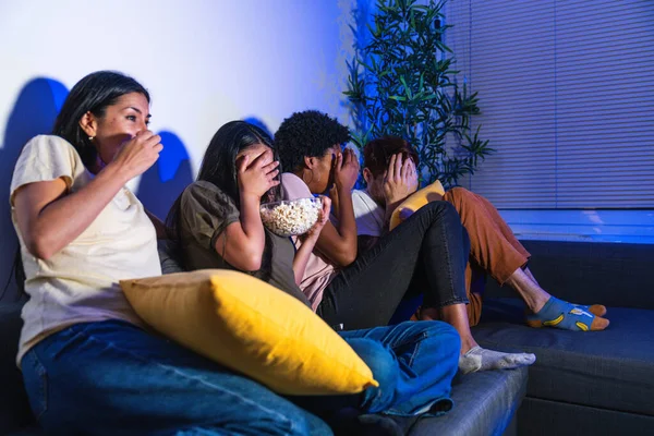 Group of diverse friends on couch at home, expressions of fear and shock while watching a horror movie, covering faces.