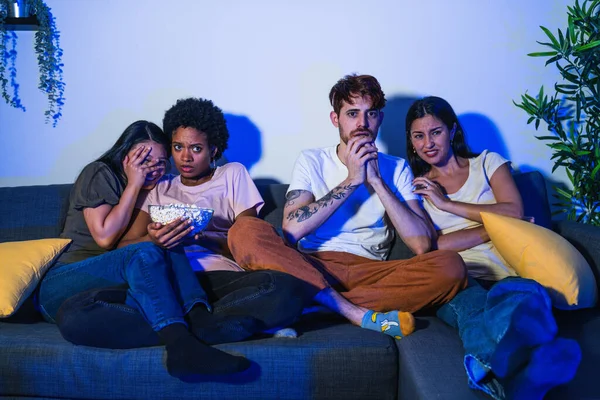 Group of friends with varied ethnic backgrounds feeling shock while watching a thrilling movie scene, expressing fear.