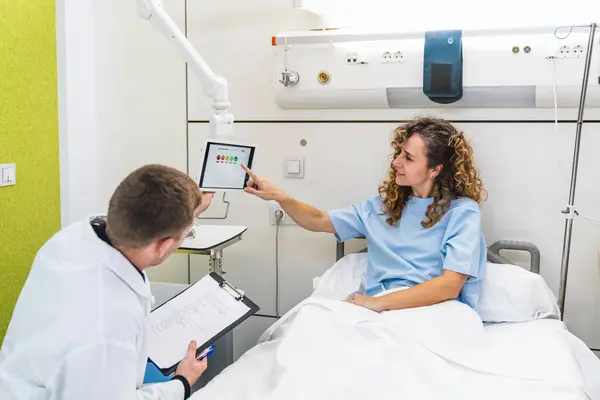 A female patient in a hospital bed interacts with a doctor by pointing at a satisfaction survey on a tablet.