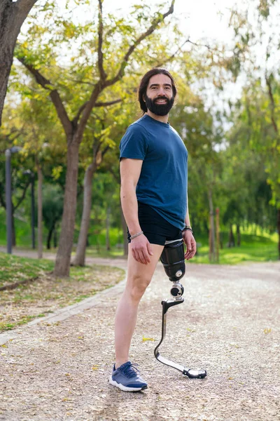Cheerful runner with an athletic leg prosthesis standing on a path, surrounded by trees.