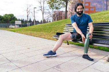 Mutlu bir sporcu parktaki bir bankta rahat bir şekilde oturur, karbon fiber protez bacağı yaklaşan fiziksel meydan okumaya hazırdır..