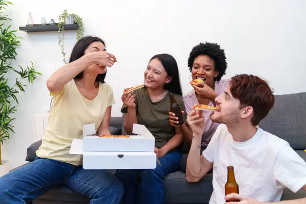 Happy young adults of diverse backgrounds enjoy pizza and beer together, laughing and relaxing on a sofa at a cozy birthday party.