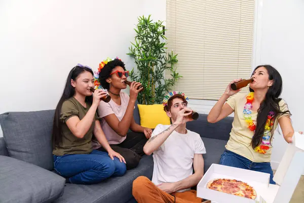 Multicultural friends eating pizza and toasting with beers on a sofa at a relaxed birthday celebration in a cozy apartment setting