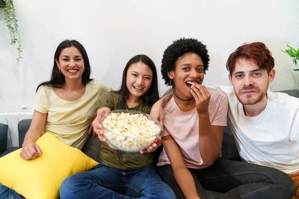 Birlikte film izleyen neşeli, çok kültürlü arkadaşlar. Gülümseyen, parlak sarı yastıklı rahat bir kanepede patlamış mısır paylaşan