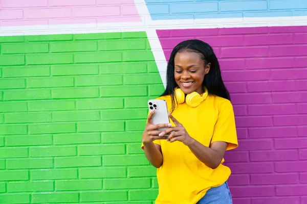 A cheerful African lady in a bright yellow top smiles at her phone, against a colorful geometric background.